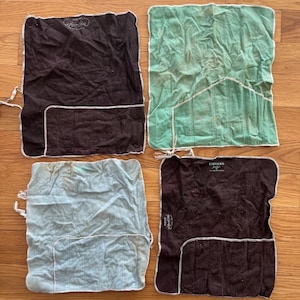 May include: Four cloth bags in shades of brown, green, and blue-green, arranged on a wooden surface. Each bag has a white stitched border and a pocket. Some bags have text printed on them.