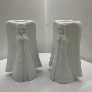 May include: Two white ceramic angel candle holders. Each angel has detailed wings and is depicted in a praying pose. The candle holders are designed with a hollowed-out top for candles.