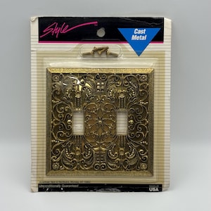 May include: A gold-colored cast metal double toggle light switch plate with an intricate embossed floral and scrollwork pattern. The product is packaged in a clear plastic blister pack on a cardboard backing labeled Style and Cast Metal.