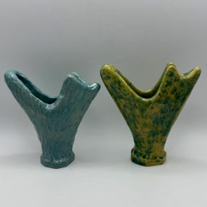 May include: Two ceramic vases with a V-shaped opening and a textured surface. One vase is a solid matte blue with a vertical ribbed pattern, while the other is a yellow base with green speckles and a smooth, glossy finish on the exterior.