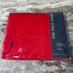 May include: A red flat sheet from Ralph Lauren, packaged in clear plastic. The sheet has a navy blue embroidered logo and a navy blue label that reads "THE POLO SHEET". The label also states the sheet is a twin size.