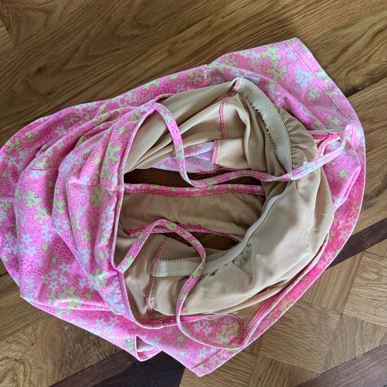 K&ouml;nnte beinhalten: Ein pinkfarbener Badeanzug mit einem Blumenmuster in Hellgr&uuml;n und Wei&szlig;. Der Badeanzug hat d&uuml;nne Tr&auml;ger und ein beiges Futter. Das Kleidungsst&uuml;ck ist gefaltet und zeigt das Innenfutter und das florale &Auml;u&szlig;ere.