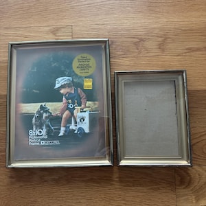 Puede incluir: Dos marcos de fotos rectangulares dorados. Uno contiene un retrato de 20 x 25 cm con un niño y un perro. El otro marco está vacío. Los marcos están sobre una superficie de madera.