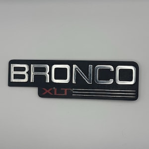 May include: A black rectangular emblem with the word "BRONCO" in silver block letters. Below the word "BRONCO" is the word "XLT" in red letters. The emblem has three horizontal silver lines.