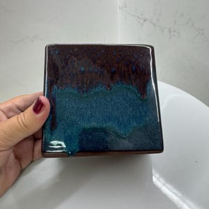 Puede incluir: Azulejo cerámico cuadrado con acabado brillante. La mitad superior es de color marrón oscuro, que pasa a un azul verdoso en la parte inferior. Los bordes son de color marrón oscuro.