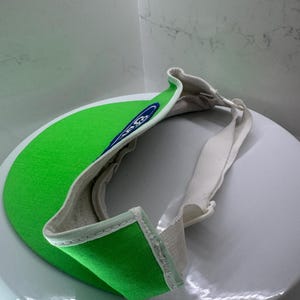 May include: A green visor with a white brim and a white adjustable strap. The visor has a blue and white logo on the front. The visor is made of a soft, textured material.