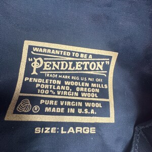 Op de afbeelding: Een donkerblauw kledingstuk van stof met een rechthoekig Pendleton-label met witte tekst: WARRANTED TO BE A PENDLETON, PENDLETON WOOLEN MILLS PORTLAND, OREGON, 100% VIRGIN WOOL, PURE VIRGIN WOOL, MADE IN U.S.A., en SIZE: LARGE.