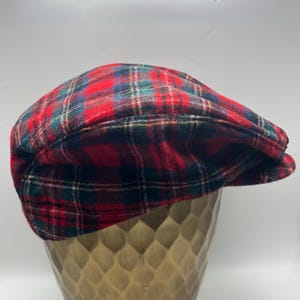 Op de afbeelding: Een platte pet met een klassiek rood, groen en blauw ruitpatroon. De pet heeft een afgeronde bovenkant en een korte, stijve klep aan de voorzijde, vervaardigd uit een zachte, geweven stof met een duidelijk zichtbare textuur in het materiaal.