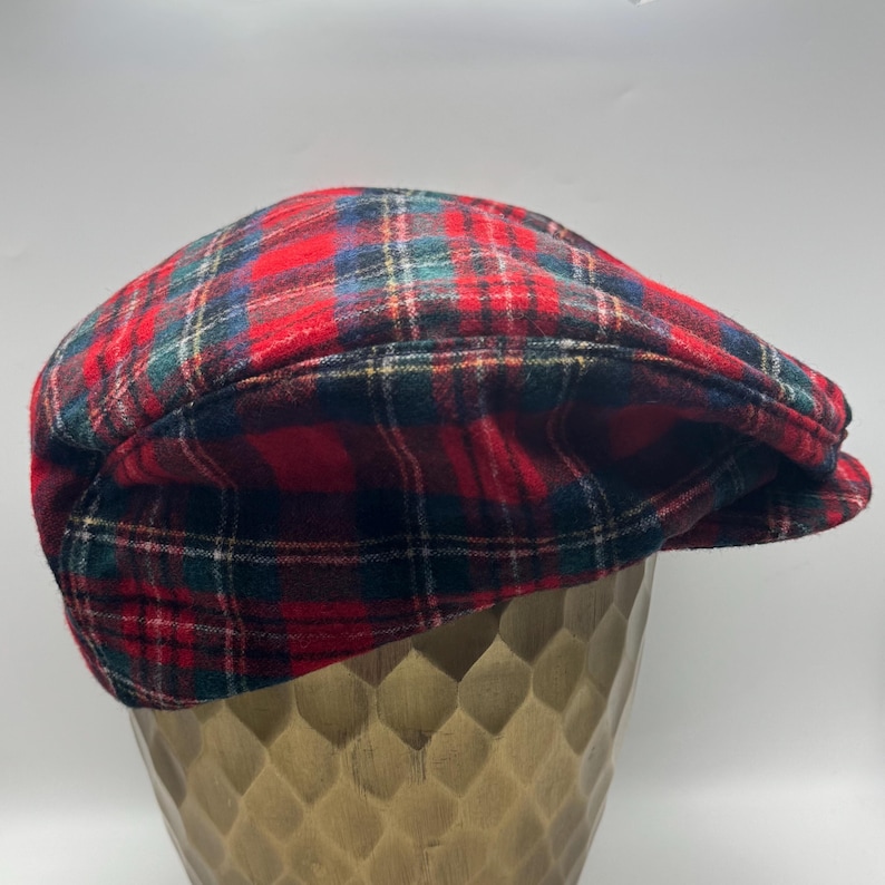 Op de afbeelding: Een platte pet met een klassiek rood, groen en blauw ruitpatroon. De pet heeft een afgeronde bovenkant en een korte, stijve klep aan de voorzijde, vervaardigd uit een zachte, geweven stof met een duidelijk zichtbare textuur in het materiaal.