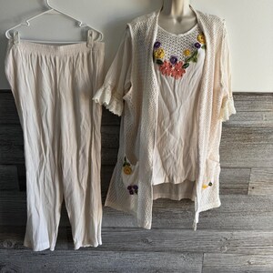 May include: A cream-colored three-piece outfit. The set includes a pair of pants, a sleeveless top, and a crocheted cardigan. The cardigan and top are adorned with floral embroidery in shades of yellow, orange, and purple. The outfit is displayed on a wooden background.