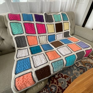Può includere: Una coperta all'uncinetto fatta a mano con un design patchwork. La coperta presenta quadrati in vari colori, tra cui rosa, blu, verde e marrone, bordati di bianco e delineati in marrone scuro. La coperta è drappeggiata su un divano grigio.