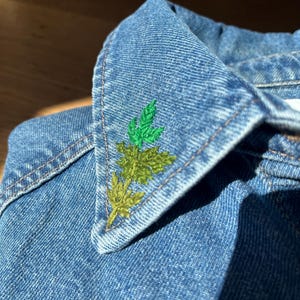 May include: Close-up of a denim jacket collar with embroidered green and yellow leaves. The collar is a light blue denim with visible stitching. The embroidery adds a touch of nature-inspired detail to the garment.