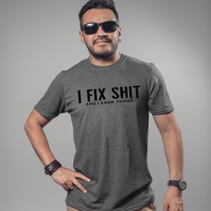 May include: A dark gray t-shirt with the text "I FIX SHIT AND I KNOW THINGS" in black. The person in the image is wearing black sunglasses and a watch. The shirt is a crew neck style.