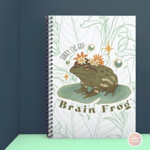 May include: A white spiral bound notebook with a green frog wearing a flower crown on a lily pad. The frog is green and brown with black stripes. The text "Sorry, I've got Brain Frog" is printed on the notebook.