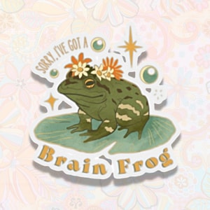 May include: A sticker featuring a green frog wearing a flower crown. The frog is sitting on a lily pad with the text "Sorry, I've got a Brain Frog" written below.