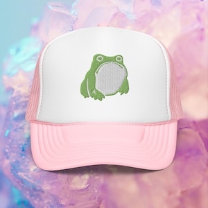May include: A pink and white trucker hat with an embroidered green frog on the front.