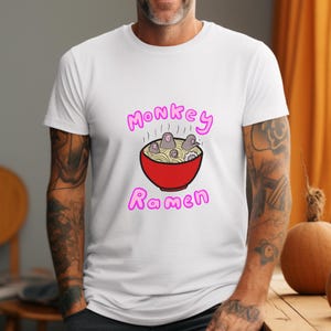 以下が含まれることがあります： 赤いラーメンの丼に猿の漫画デザインが施された白いTシャツ。「Monkey Ramen」の文字がピンク色で丼の上と下に書かれています。このTシャツは柔らかく快適な素材で作られています。