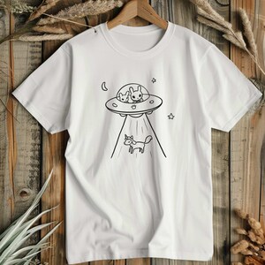 Könnte beinhalten: Ein weißes Kurzarm-T-Shirt mit einer schwarzen Strichzeichnung einer fliegenden Untertasse, in der zwei hasenartige Wesen sitzen und ein kleines Einhorn mit einem Lichtstrahl nach unten befördern. Das Motiv zeigt zudem eine Mondsichel und zwei Sterne.