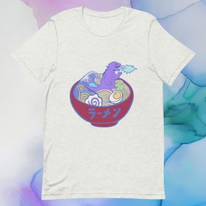 May include: A light grey t-shirt with a graphic of a purple Godzilla monster in a bowl of ramen. The ramen bowl is red with the Japanese word "Ramen" written on it. The ramen bowl is filled with noodles, an egg, seaweed, and a wave.