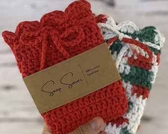 Könnte beinhalten: Zwei handgestrickte Seifensäckchen aus Baumwolle. Eines ist einfarbig rot, das andere ist weiß mit roten und grünen Akzenten. Jedes hat einen Kordelzugverschluss und einen gewellten Rand. Ein braunes Etikett trägt die Aufschrift "Soap Saver" und "100% cotton hand-knit".