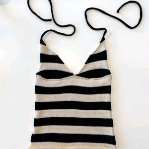 May include: A sleeveless halter top featuring a horizontal striped pattern in black and beige. The garment is constructed with a V-neckline and includes two long black knitted ties at the top for fastening behind the neck, creating a simple silhouette.