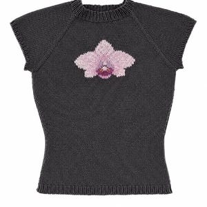 May include: A dark charcoal gray short-sleeved knit top featuring a ribbed crew neckline and hem. The center of the chest displays a detailed intarsia knit graphic of a light pink orchid flower with a deep purple and white center detail.