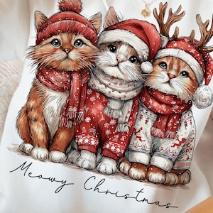 May include: White sweatshirt featuring a graphic of three festive kittens wearing Santa hats, scarves, and sweaters. The kittens are in shades of orange, white, and red. The text "Meowy Christmas" is written in a script font below the kittens.