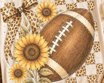 May include: A cream-colored short-sleeve t-shirt featuring a graphic print of a brown football, three yellow sunflowers with green leaves, a large leopard print bow, and a leopard print checkered pattern background across the chest area.
