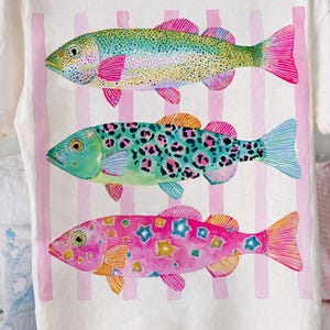 May include: White t-shirt featuring three colorful fish designs. The fish have vibrant patterns and colors, including pink, blue, green, and yellow. Vertical pink stripes are behind the fish. The shirt is held up by a person.