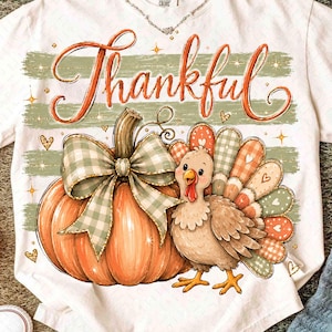 May include: A white short-sleeve t-shirt featuring a graphic of an orange pumpkin with a green and white checkered bow, a brown turkey, and the word Thankful in orange script. The design includes green horizontal stripes and small gold stars.