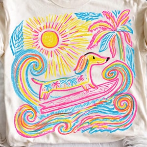 May include: A cream-colored t-shirt featuring a vibrant graphic print of a yellow dachshund surfing on a pink surfboard. The design includes a bright yellow sun, a pink palm tree, and swirling blue and pink ocean waves surrounding the dog.