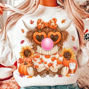 May include: A white long-sleeve sweatshirt featuring a graphic of a fluffy highland cow wearing a pumpkin-patterned bow, heart-shaped pumpkin glasses, and blowing a pink bubble. The cow wears a cream sweater and sits among sunflowers and pumpkins.
