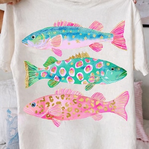 May include: A white t-shirt featuring three colorful fish illustrations. The fish have blue, green, and pink bodies with gold accents. The shirt is held up by a person.