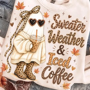 Könnte beinhalten: Ein weißes Sweatshirt mit einer Grafik eines Geistes, der einen cremefarbenen Pullover, Leoparden-Stiefel und herzförmige Sonnenbrillen trägt und einen Eiskaffee hält. Der Text lautet Sweater Weather & Iced Coffee, umgeben von Ahornblättern.
