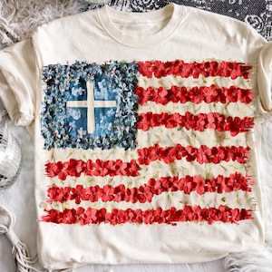 Puede incluir: Camiseta de color crema con un diseño floral de la bandera estadounidense. Las rayas de la bandera están hechas de flores rojas, el cuadrado azul está lleno de flores azules y una cruz blanca está en el centro. La camiseta tiene cuello redondo y mangas largas.
