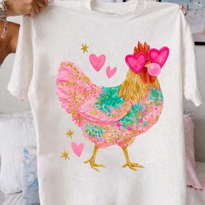 May include: White t-shirt featuring a colorful chicken graphic. The chicken has pink and teal feathers with gold accents, heart-shaped sunglasses, and a bubblegum bubble. Pink hearts and gold stars are also printed on the shirt.