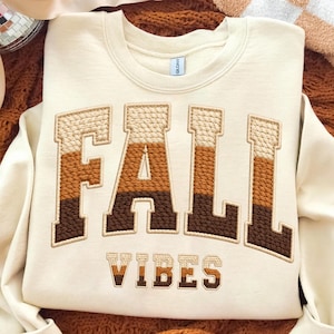 May include: A cream-coloured crewneck sweatshirt featuring the word FALL in large, block-style embroidered letters with a three-tone gradient of beige, tan, and dark brown. Below, the word VIBES is embroidered in a smaller, matching brown font.