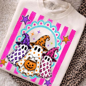 May include: A cream-colored t-shirt featuring a graphic of three ghosts wearing witch hats. The ghosts are decorated with purple, orange, and pink patterns, and one holds a jack-o'-lantern. The design includes pink stripes and stars.