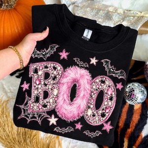 Op de afbeelding: Een zwart sweatshirt met ronde hals en het woord BOO in grote letters. De B en de O zijn versierd met roze en zwarte strass-steentjes, terwijl de middelste O is bedekt met roze imitatiebont. Het ontwerp bevat roze sterren, zwarte vleermuizen en een spinnenweb.