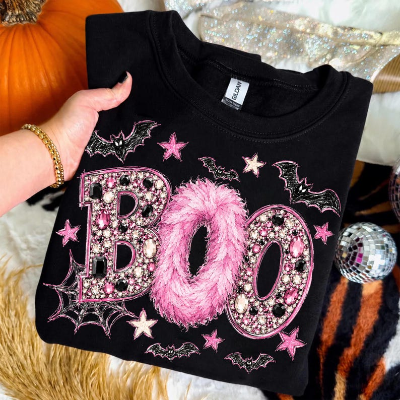 Op de afbeelding: Een zwart sweatshirt met ronde hals en het woord BOO in grote letters. De B en de O zijn versierd met roze en zwarte strass-steentjes, terwijl de middelste O is bedekt met roze imitatiebont. Het ontwerp bevat roze sterren, zwarte vleermuizen en een spinnenweb.