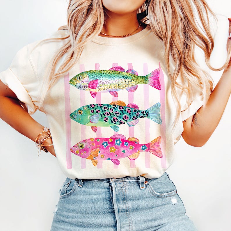 May include: Cream-colored t-shirt featuring three colorful fish illustrations. The fish have pink fins and are set against a backdrop of pink vertical stripes. The fish designs include a rainbow, leopard print, and floral patterns.