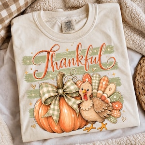 May include: A cream-colored t-shirt featuring a graphic of an orange pumpkin with a green gingham bow and a cartoon turkey with patterned tail feathers. The word Thankful is printed in orange script above the pumpkin, accented by small gold stars.
