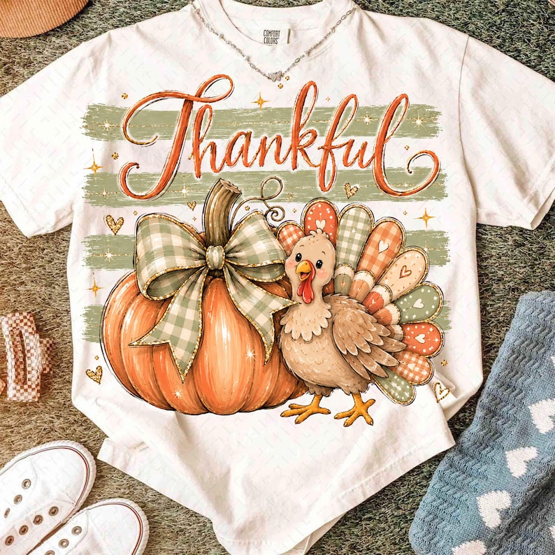 May include: A white short-sleeve t-shirt featuring a graphic of an orange pumpkin with a green and white checkered bow, a brown turkey, and the word Thankful in orange script. The design includes green horizontal stripes and small gold stars.