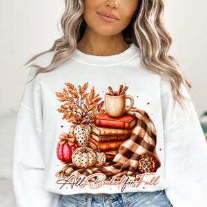 Peut inclure: Un sweat-shirt blanc &agrave; col rond orn&eacute; d'une illustration repr&eacute;sentant une pile de livres, une tasse avec des b&acirc;tons de cannelle, des feuilles d'automne et des citrouilles. Le motif inclut une couverture &agrave; carreaux et le texte "All Booked for Fall".