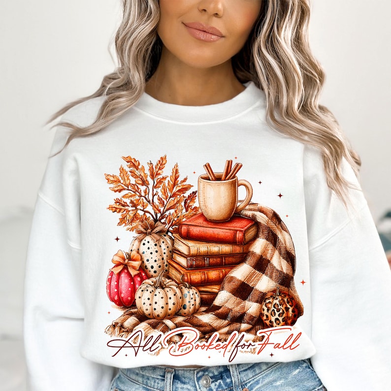 Peut inclure: Un sweat-shirt blanc &agrave; col rond orn&eacute; d'une illustration repr&eacute;sentant une pile de livres, une tasse avec des b&acirc;tons de cannelle, des feuilles d'automne et des citrouilles. Le motif inclut une couverture &agrave; carreaux et le texte "All Booked for Fall".
