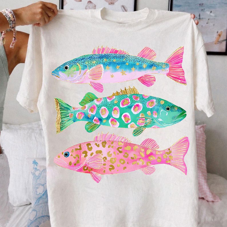 May include: A white t-shirt featuring three colorful fish illustrations. The fish have blue, green, and pink bodies with gold accents. The shirt is held up by a person.