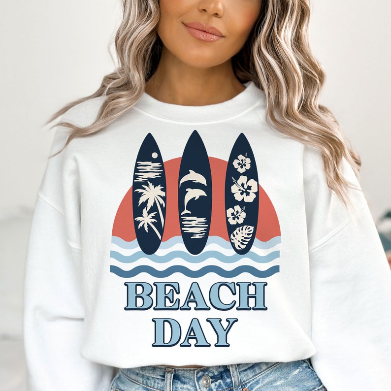 May include: White sweatshirt with a graphic of three surfboards with palm trees, dolphins, and hibiscus flowers. The graphic is set against a red sunset and blue waves. The text "BEACH DAY" is printed below the graphic.
