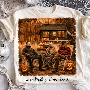 May include: A cream-colored t-shirt featuring a graphic of three horror movie characters sitting in lawn chairs in front of a cabin at night. The scene includes lit jack-o'-lanterns and the text "mentally i'm here" printed in black script.
