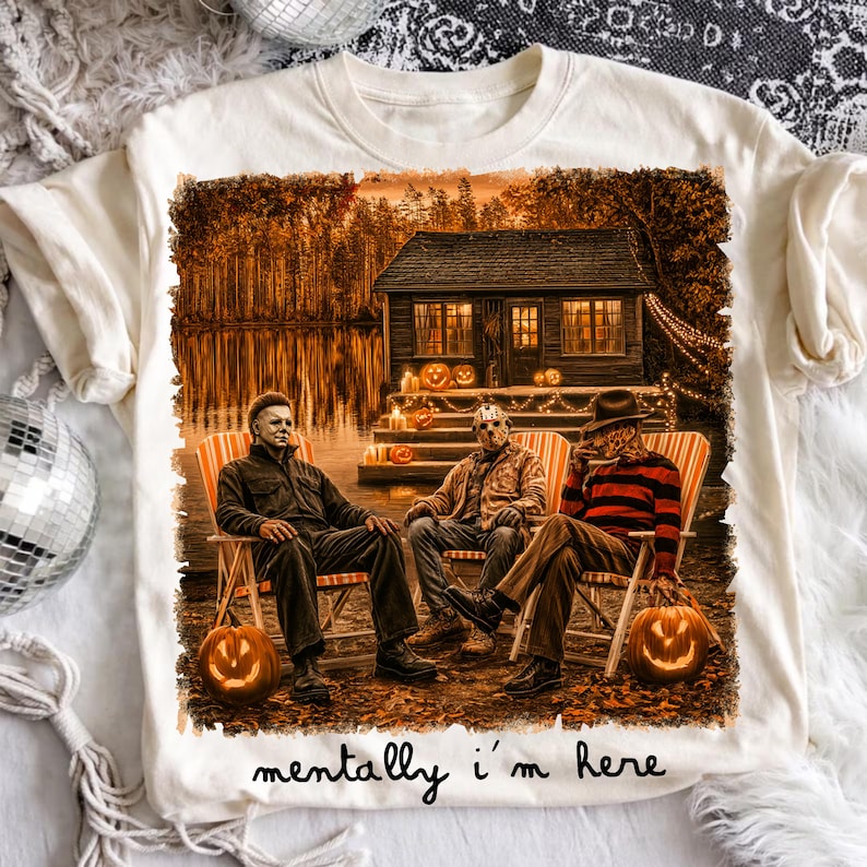 May include: A cream-colored t-shirt featuring a graphic of three horror movie characters sitting in lawn chairs in front of a cabin at night. The scene includes lit jack-o'-lanterns and the text "mentally i'm here" printed in black script.