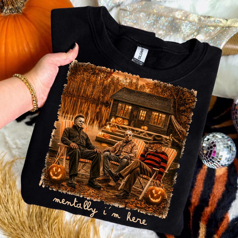 May include: A black sweatshirt featuring a graphic print of three horror movie characters sitting in chairs outside a cabin at night. The print includes pumpkins and the text "mentally i'm here" in white script lettering below the illustration.